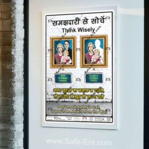 A 'Think Wisely' behaviour safety poster (Code: BSP-6) framed and displayed on an office wall. The poster contrasts two framed pictures: the left shows a 'Safe & Happy Family,' while the right shows an 'Unsafe & Unhappy Family,' with the father's silhouette missing from the frame. The message reads, 'Don't mock Safety's Call, or you'll be Absent from the Wall' in English and a similar warning in Hindi.